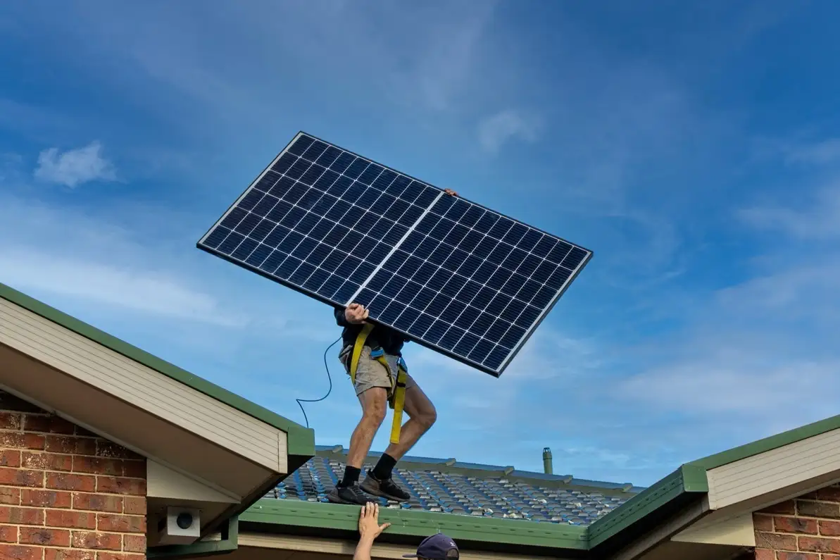 A person in a harness standing on a tiled roof holding a solar panel above their head against a blue sky, with another person's hands reaching up from the gutter below