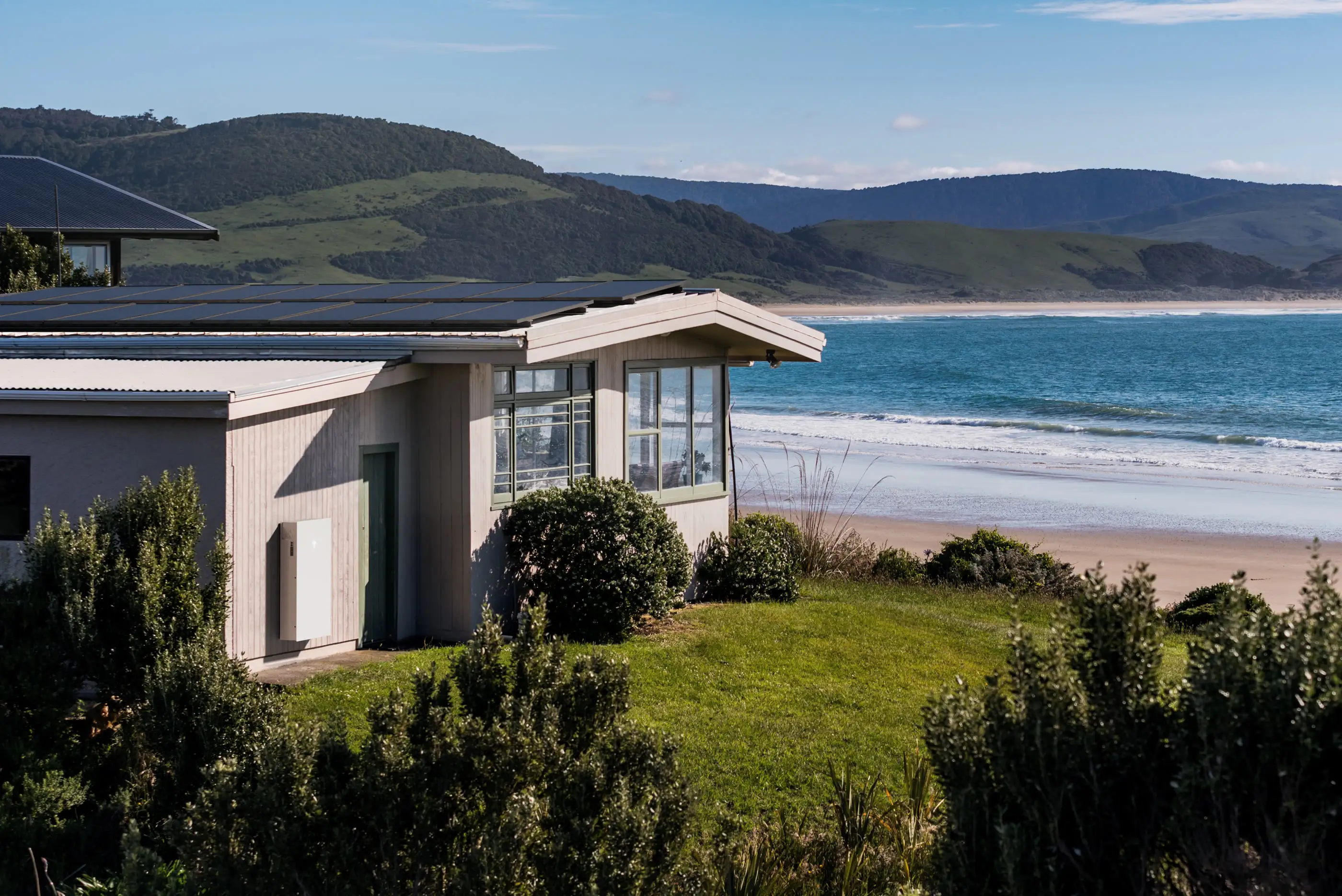 A low weatherboard house with black solar panels running the full length of its shallow metal roof and a white home battery mounted on the wall beside the door, lawn and dune scrub in front of it, surf and green headlands behind