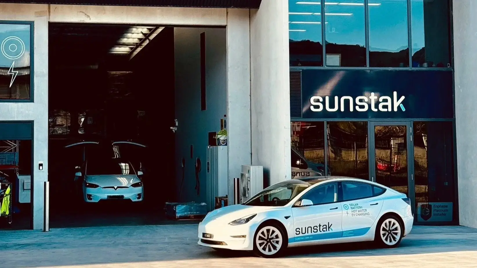 Two Teslas at the Sunstak premises in Hume: a white Model 3 in Sunstak livery parked in front of the glass shopfront, and a white Model X with its doors raised inside the open roller door beside it