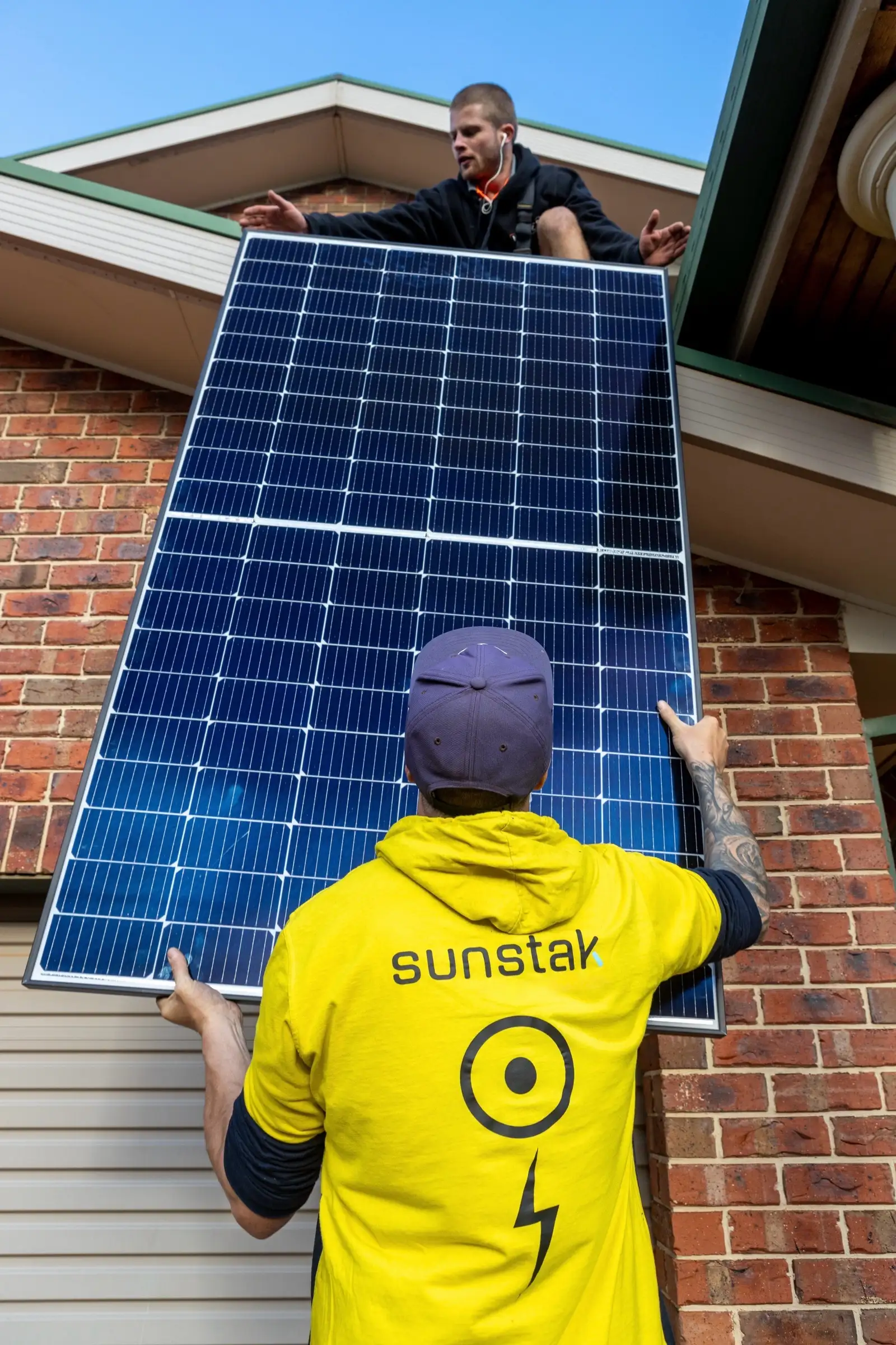 Two people passing a solar panel up the brick wall of a house, seen from below: one in a yellow Sunstak hoodie and cap holding it from underneath with both hands, the other leaning down over the eaves in a dark jacket, against a blue sky
