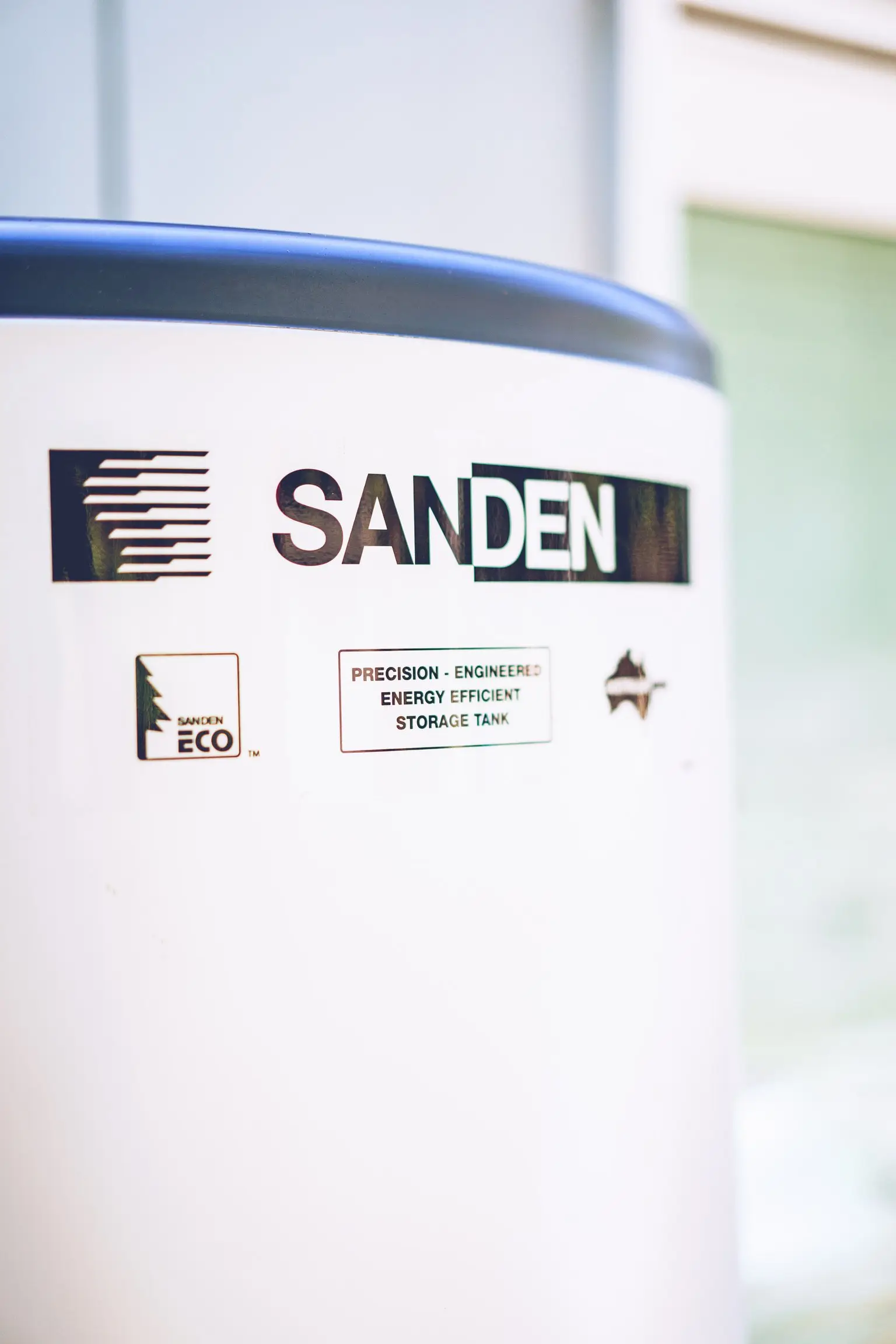 Close-up of the Sanden nameplate on a stainless steel hot water tank, reading Precision-Engineered Energy Efficient Storage Tank.