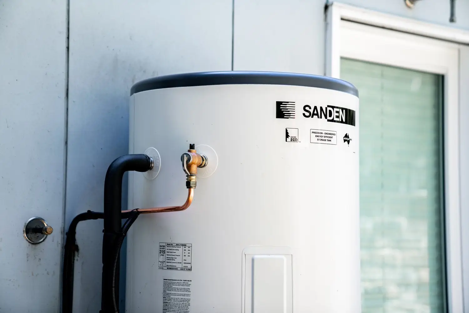 A stainless steel hot water tank branded Sanden, installed against an exterior house wall with copper and PVC pipework connected at its base.