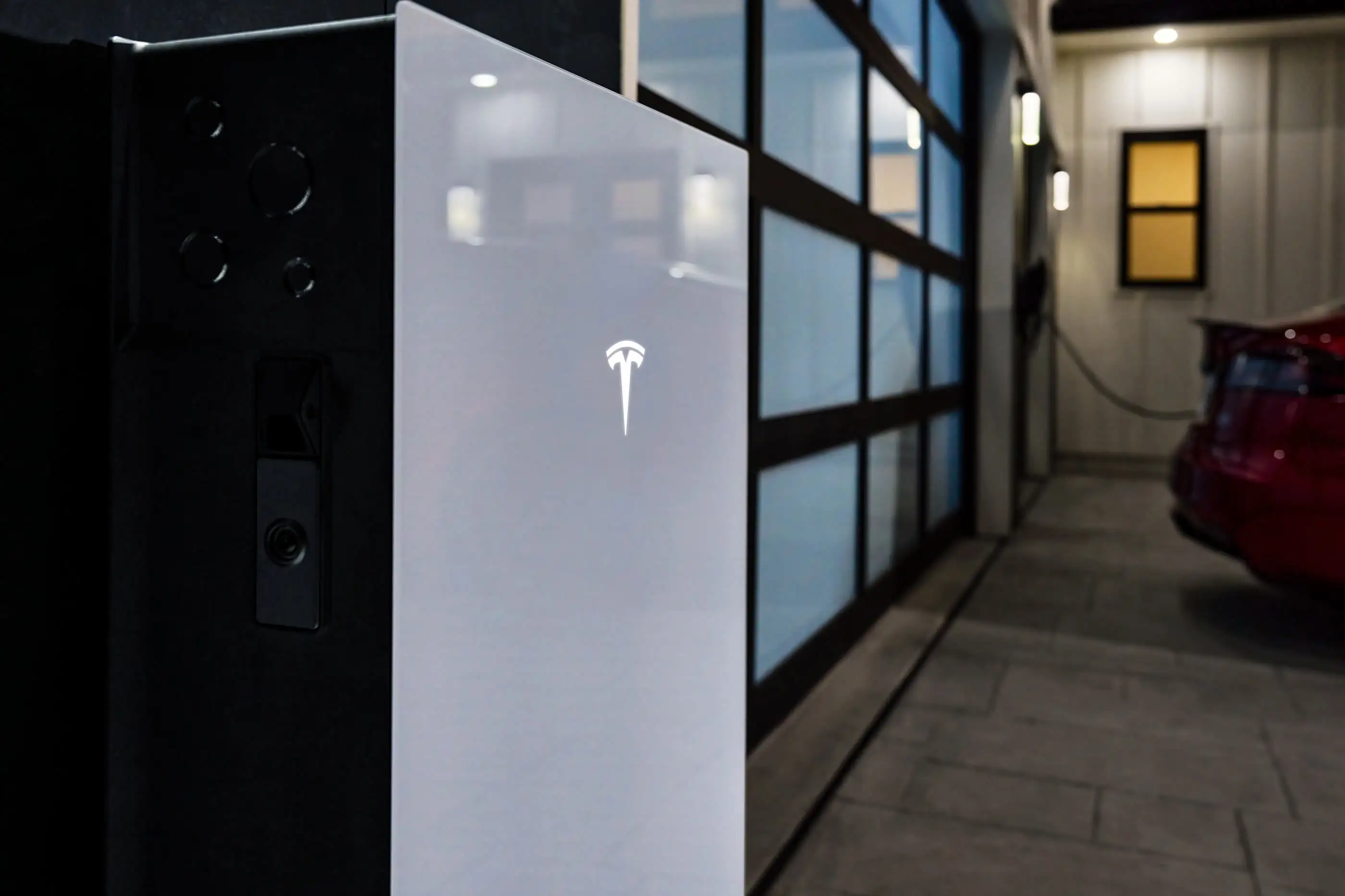 A white home battery seen close up from one side, a lit Tesla mark on its face, with its dark side panel, four blanked knockouts and a black service fitting to the left, and a black-framed glass wall behind it